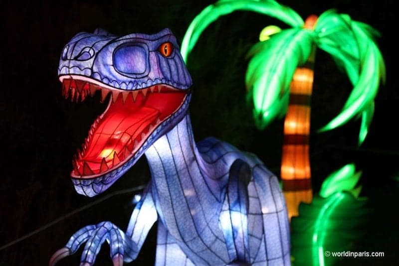 Aquí están las mejores ciudades de Europa en invierno para familias, dinosaurio raptor hecho de papel translúcido iluminado desde dentro con luces moradas y rojas de pie frente a otra estructura iluminada con la forma y los colores de una palmera