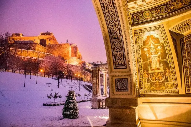 Vea las ciudades europeas más hermosas desde arriba este invierno, estructura de piedra tallada y pintada ornamentada con vista a un área de parque detrás con un alto edificio de castillo fortificado y árboles cubiertos de nieve