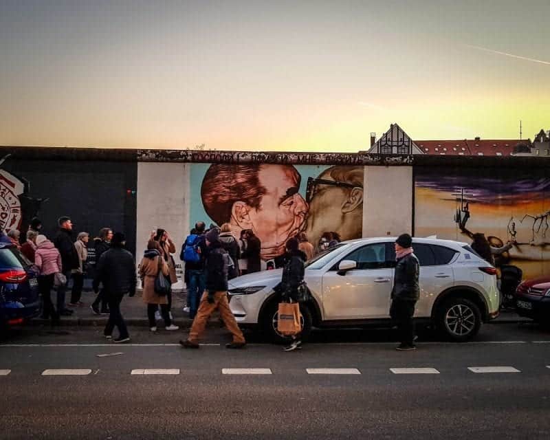Encuentre las mejores ciudades europeas en invierno, vista de la calle junto al Muro de Berlín con arte callejero elaborado con peatones tomando fotografías junto a autos estacionados al atardecer