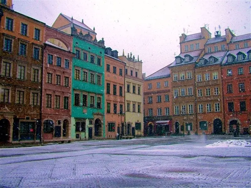 Explore Europa para conocer las mejores ciudades para visitar en invierno, una gran plaza abierta con edificios de colores brillantes que rodean un área plana pavimentada cubierta con una ligera capa de nieve blanca