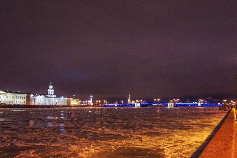Disfrute de los mejores lugares para visitar en Europa en invierno, un gran río ancho con un puente azul iluminado a lo lejos bajo un cielo nocturno nublado