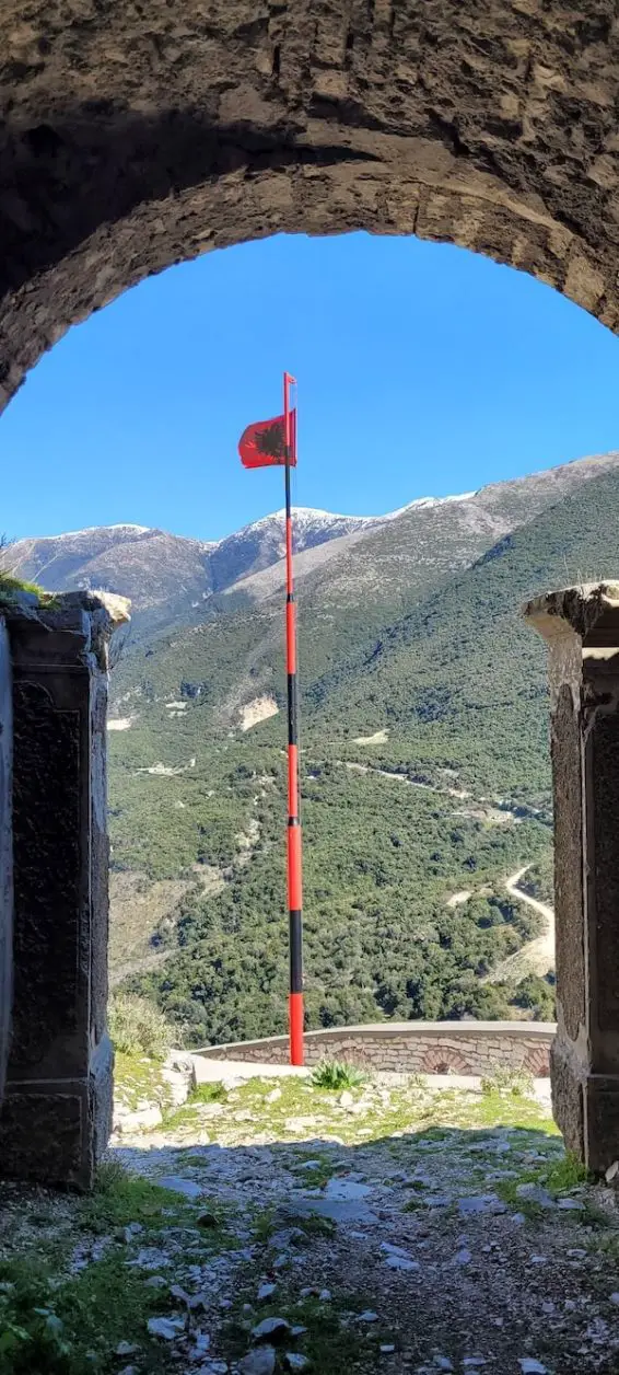 Visite los castillos de Albania, un alto asta de bandera roja y negra que muestra la bandera albanesa ondeando en el viento vista entre las fortificaciones de piedra del castillo de Borsh con montañas al fondo