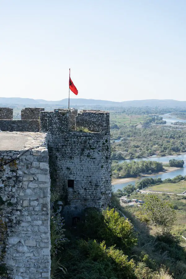 Explore los castillos en Albania, la pared de la torreta del castillo de Rozafa con la bandera roja albanesa ondeando desde el asta de la bandera sobre una gran área verde abierta que conduce hacia las montañas en la distancia