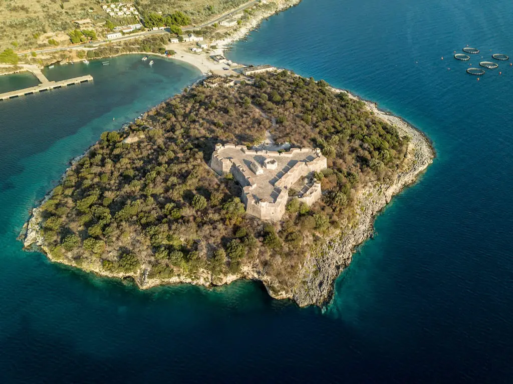 Visite el mejor castillo de Albania, el castillo de Porto Palermo visto desde arriba con muros fortificados en forma de triángulo en una pequeña isla cubierta de árboles conectada al continente por una pequeña lengua de tierra