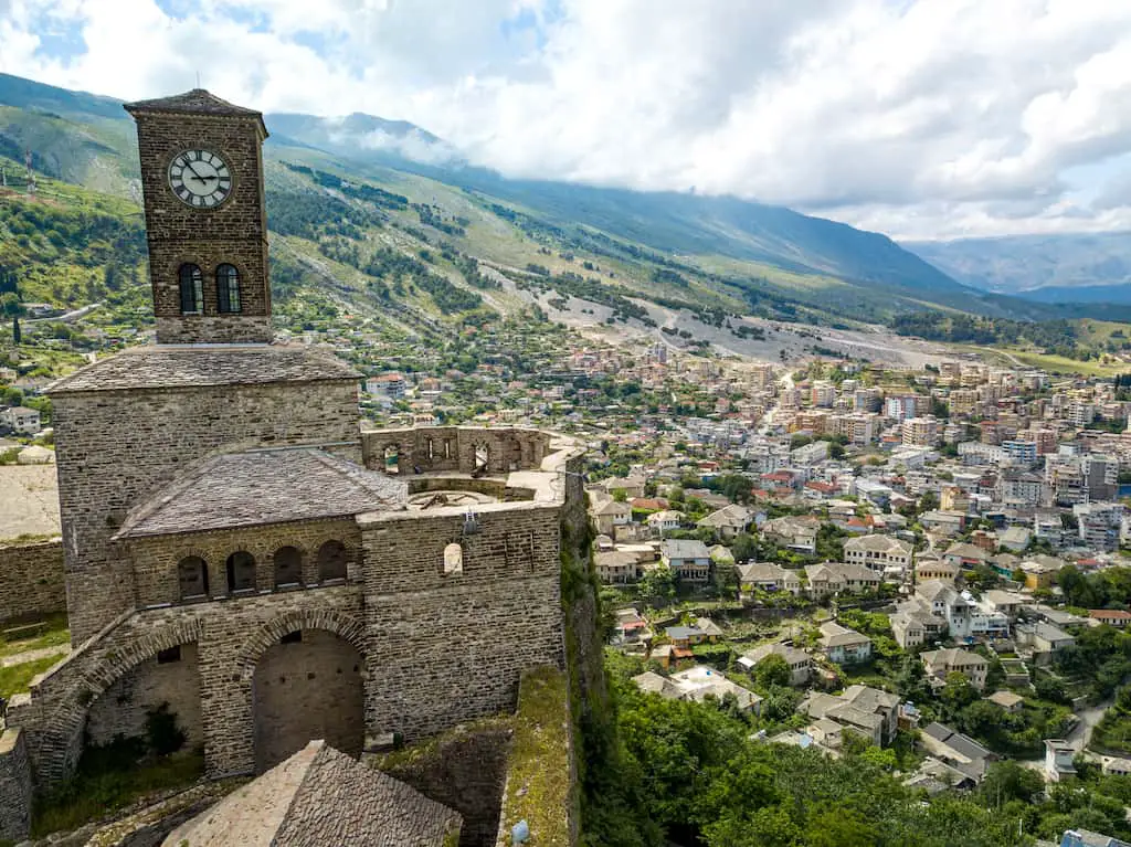 Encuentre sus castillos favoritos de Albania, el castillo de Gjirokastra con paredes de piedra y una gran torre de reloj rectangular con vista al valle con muchos edificios modernos junto a una gran ladera ondulada bajo un cielo nublado
