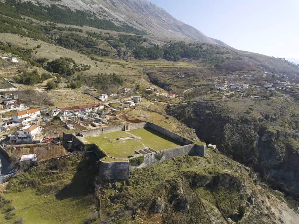 Explore todos los castillos de Albania, vista aérea del castillo de Libohovës con grandes muros de piedra en lo alto de una colina con edificios modernos cerca de una colina verde y ondulada