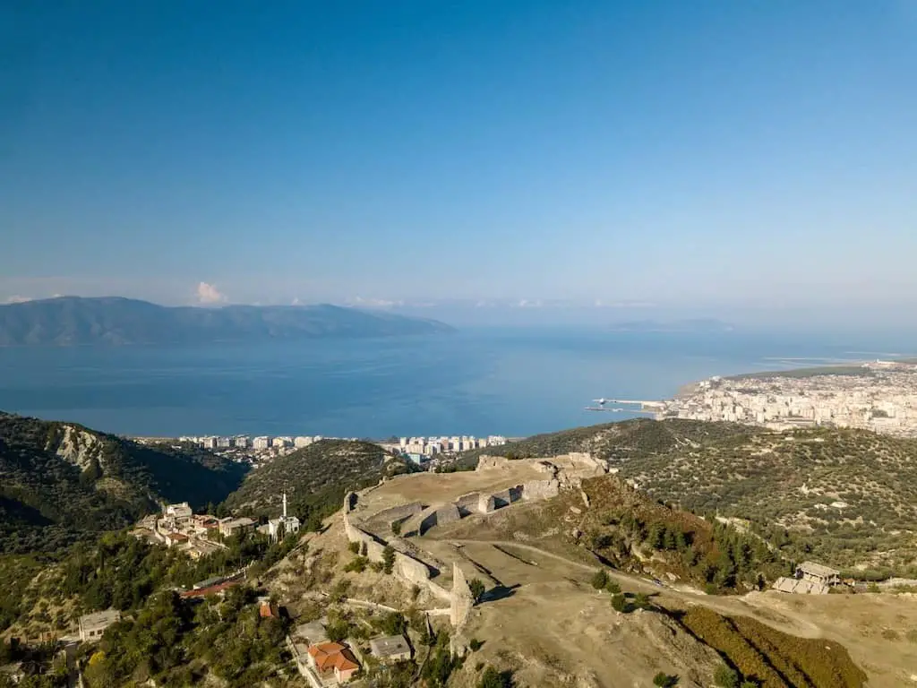 Explore los castillos de Albania, las ruinas de la ladera del castillo de Kanina con vista a la bahía junto al mar azul celeste y las áreas urbanizadas modernas más allá con las montañas en la distancia