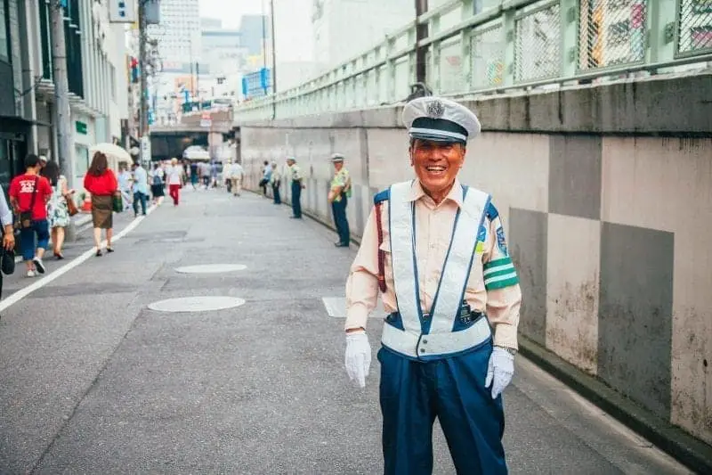 Datos curiosos de Japón, guardia de tráfico sonriente con peatones pasando junto a más guardias de tráfico calle abajo en la distancia