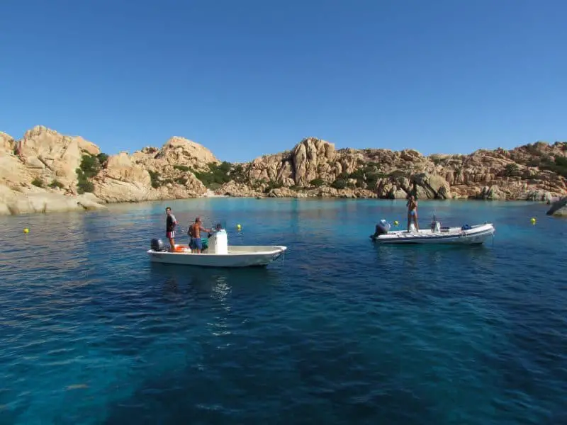 Estas son algunas de las mejores playas de Cerdeña, dos pequeños botes con personas de pie en ellos flotando en aguas profundas y ricas en azul con un anillo de tierra rocosa detrás de ellos bajo un cielo azul claro