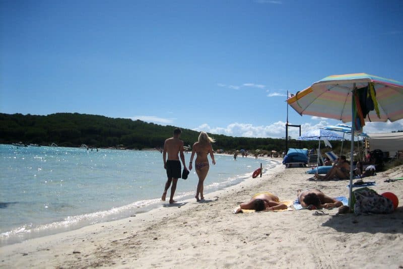 Descubre las mejores playas de Cerdeña, dos bañistas caminando por una playa de arena junto a varios bañistas y su parafernalia de playa con colinas cubiertas de árboles en la distancia bajo un cielo azul