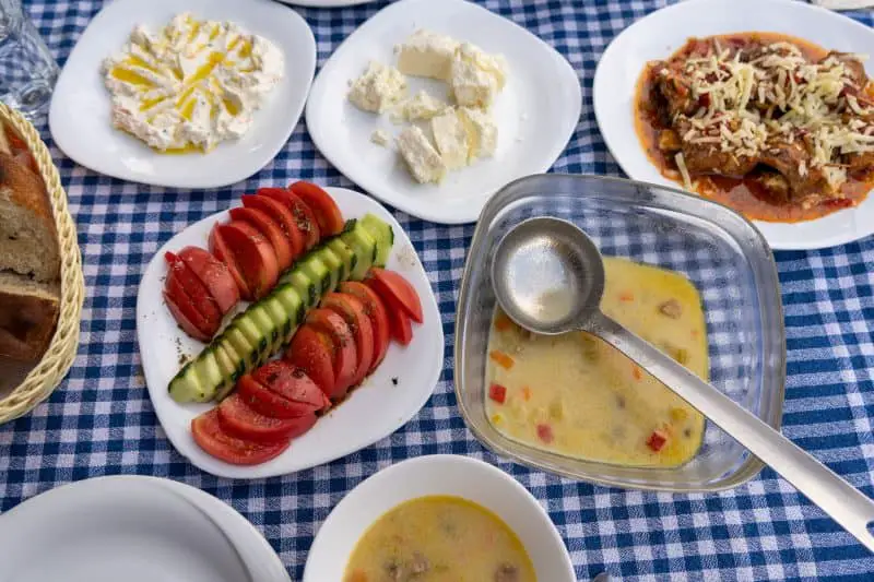Encuentre su manjar albanés favorito, varios platos de platos albaneses que incluyen carne cubierta con queso rallado y salsa, ensalada de tomate y pepino, queso feta y salsa cremosa, todo sobre un mantel azul y blanco.