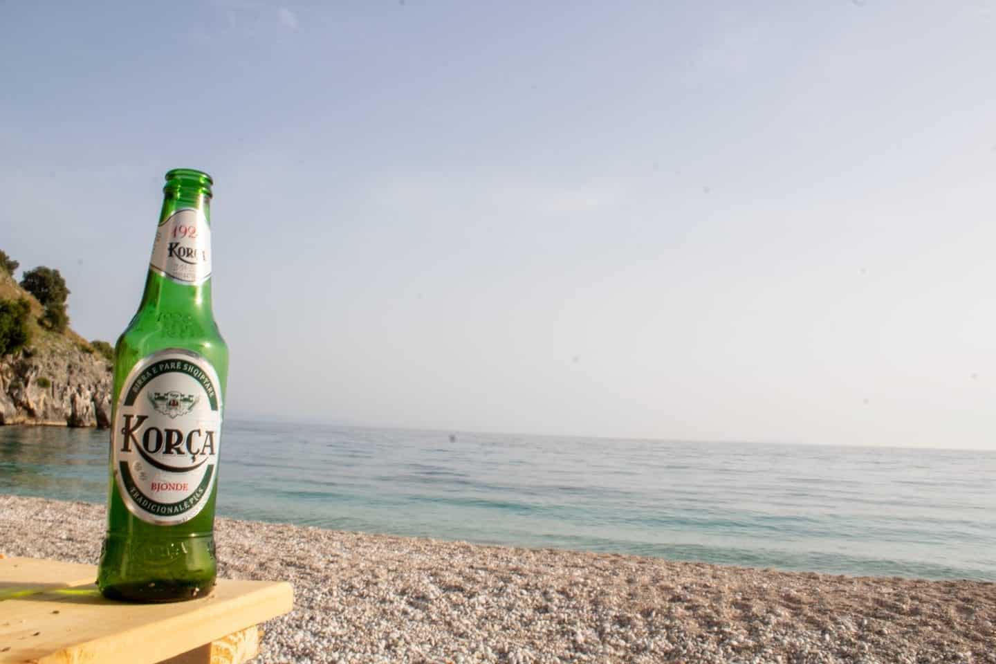 Pruebe las comidas de Albania, una botella de cerveza Korca en una mesa con una playa pedregosa y el océano al fondo