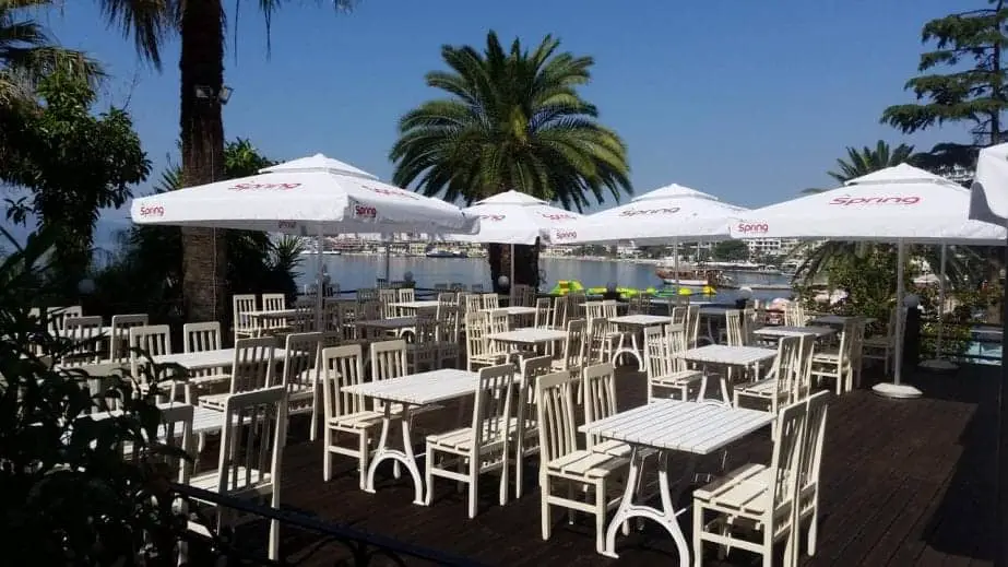 Encuentra los mejores hoteles en Saranda, zona de estar al aire libre con mesas y sillas de madera blanca con sombrillas y palmeras junto a un puerto bajo un cielo azul