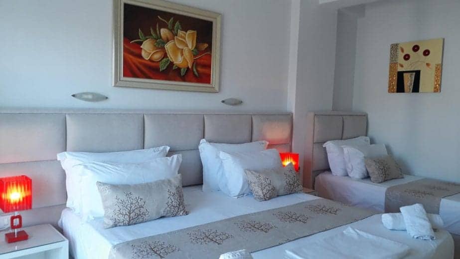 Vea el mejor hotel en Saranda, habitación de hotel con cama doble y cama individual con sábanas blancas y grises cuidadosamente preparadas, así como lámparas de mesa de color naranja brillante y cuadros de flores de arte moderno.