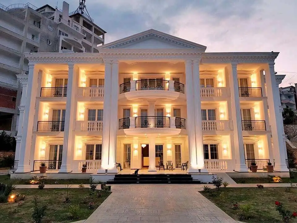Hospédese en los mejores hoteles de lujo en Saranda, frente imponente del hotel con columnas blancas y luces amarillas brillantes con balcones simétricos y área de porche
