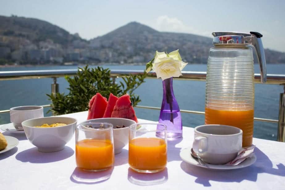 Los mejores hoteles de 5 estrellas en Albania, mesa con vasos de jugo de frutas y tazas de té con flores decorativas en un vaso, todo frente al agua con montañas a lo lejos
