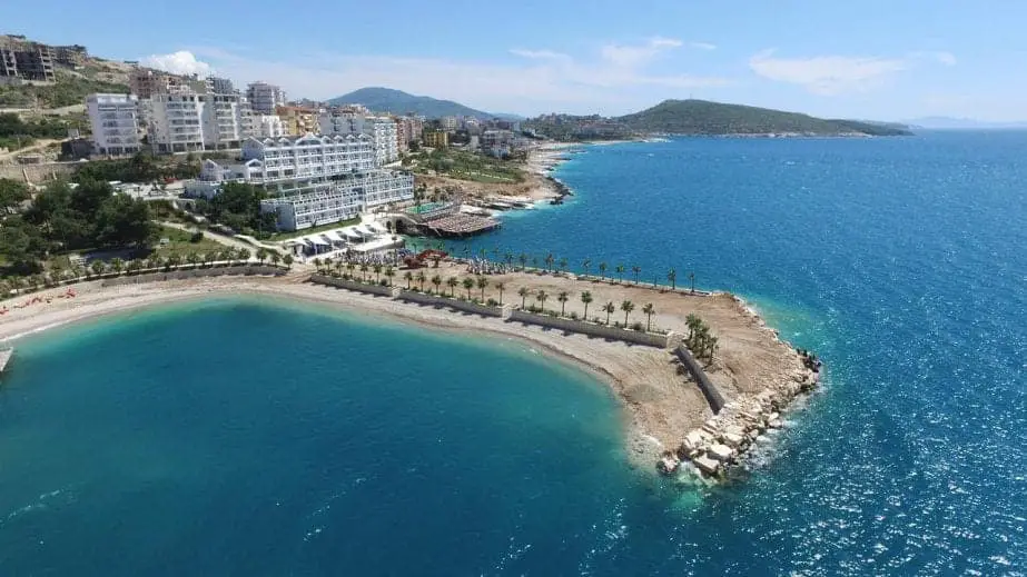 Encuentre los mejores hoteles de lujo de Albania, hotel con embarcadero de playa bordeado de palmeras rodeado de agua azul celeste con montañas en la distancia
