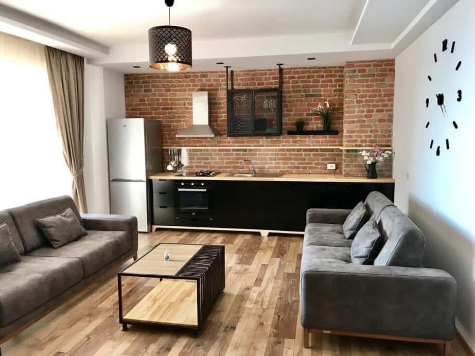 Explore dónde alojarse en Saranda Albania, un apartamento acogedor pero moderno con dos grandes sofás grises y una mesa de café con área de cocina detrás y un gran reloj en la pared