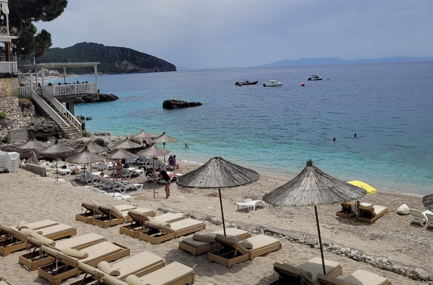 Los mejores hoteles de playa de Dhermi, sillas de playa, algunas con sombrillas de paja, en la playa rodeada de mar azul claro en un día soleado