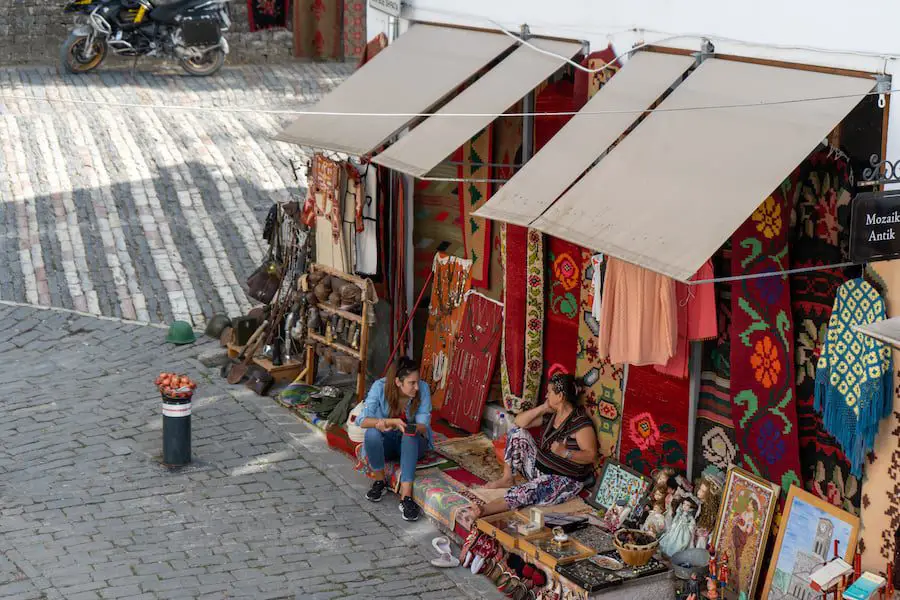 Los mejores recuerdos albaneses para comprar, dos mujeres sentadas afuera de un puesto al aire libre con muchos coloridos recuerdos albaneses