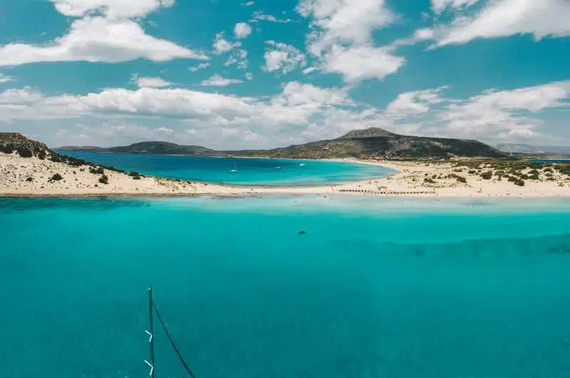 30 de los mejores lugares para visitar en Grecia, agua azul turquesa y playa de arena blanca con islas a lo lejos bajo un cielo azul con nubes