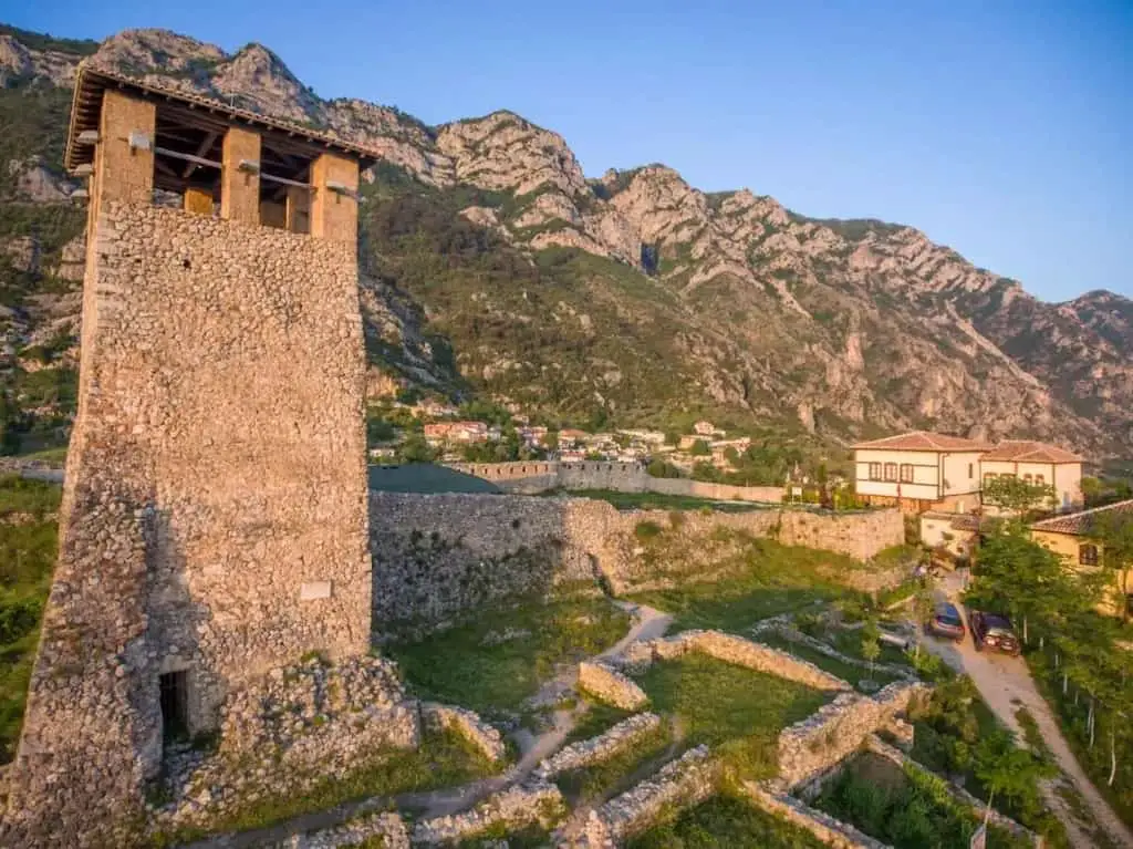 Las mejores cosas para hacer en Kruja, Albania, el castillo de Kruja con una gran torre de piedra y ruinas de pequeños muros de piedra junto a edificios modernos y una gran serie de picos montañosos bajo un cielo azul celeste