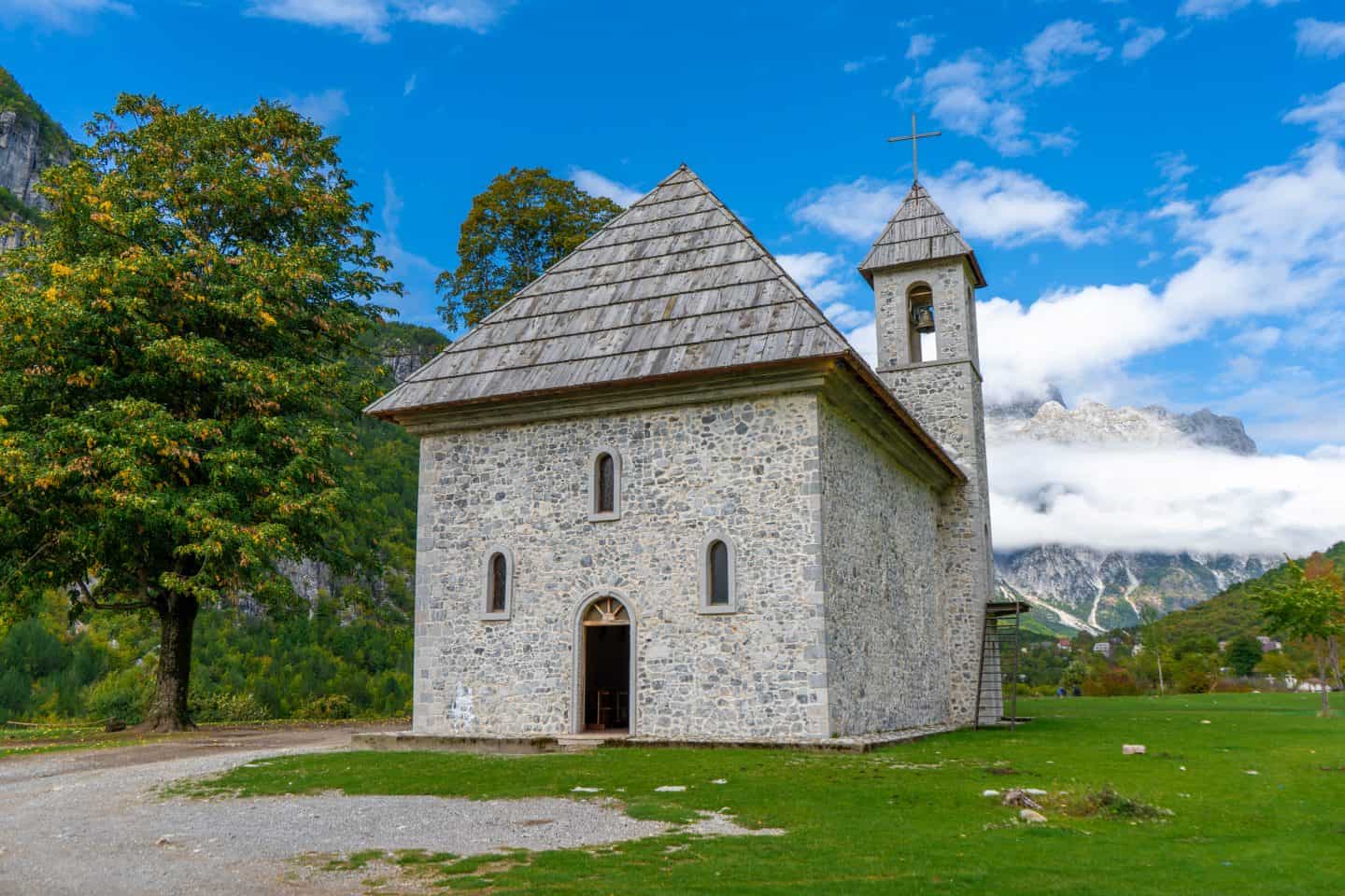 Destinos europeos baratos que debes visitar, iglesia gris con paredes de piedra, techo piramidal y torre única junto a árboles verdes con montañas empañadas en la distancia