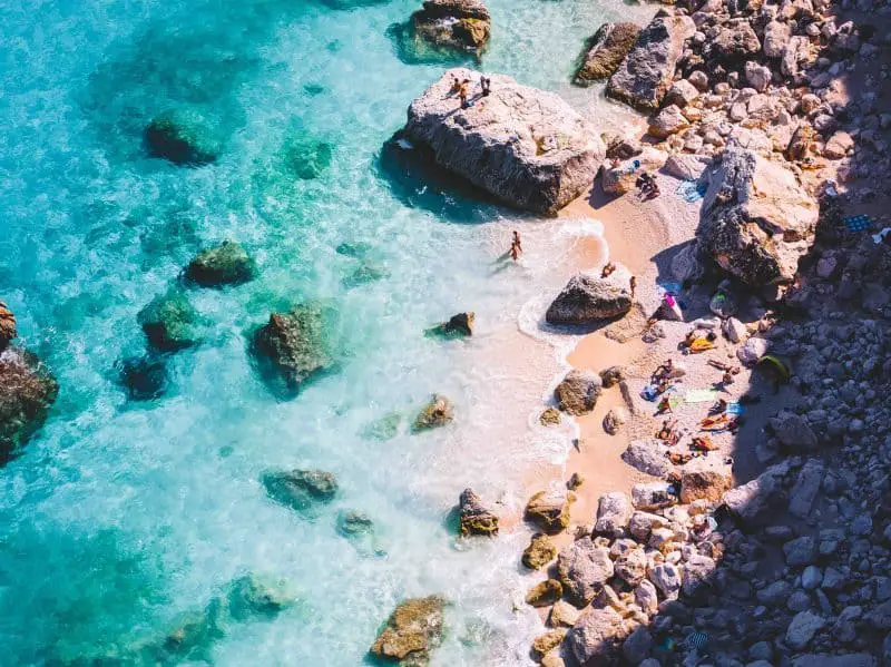 Explora las mejores playas de Cerdeña, gente tomando el sol y nadando en una playa de arena con formaciones rocosas y aguas cristalinas de color turquesa.