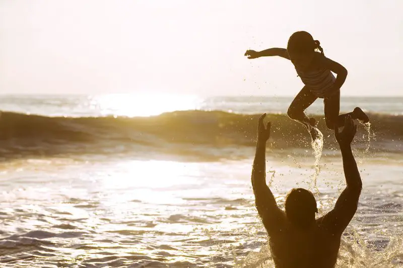 Padre jugando y arrojando a su hija en el océano al atardecer