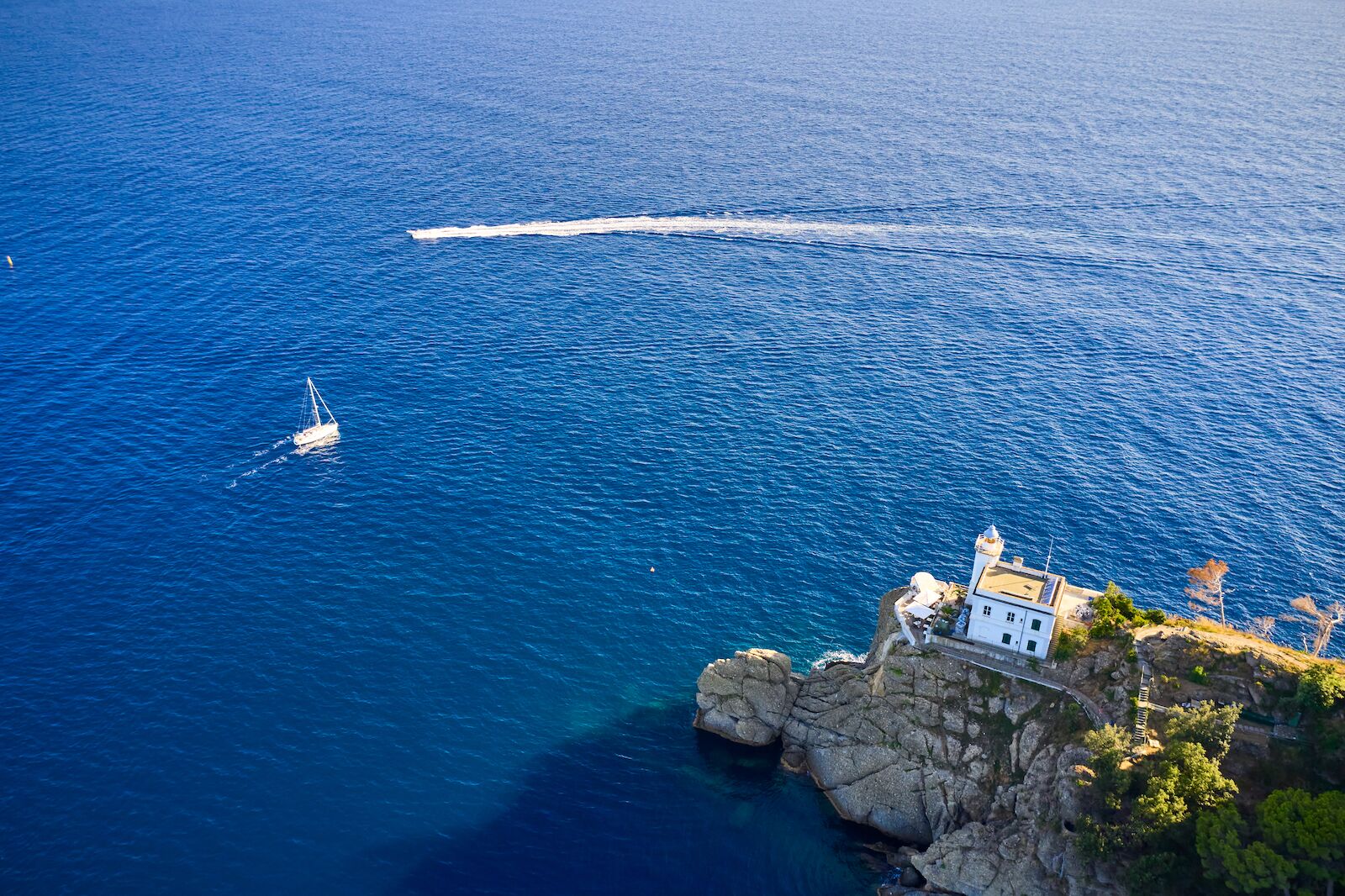 Un yate blanco y una lancha motora navegan cerca de un faro en Portofino, Italia. Un faro está situado en la colina cerca de una playa.