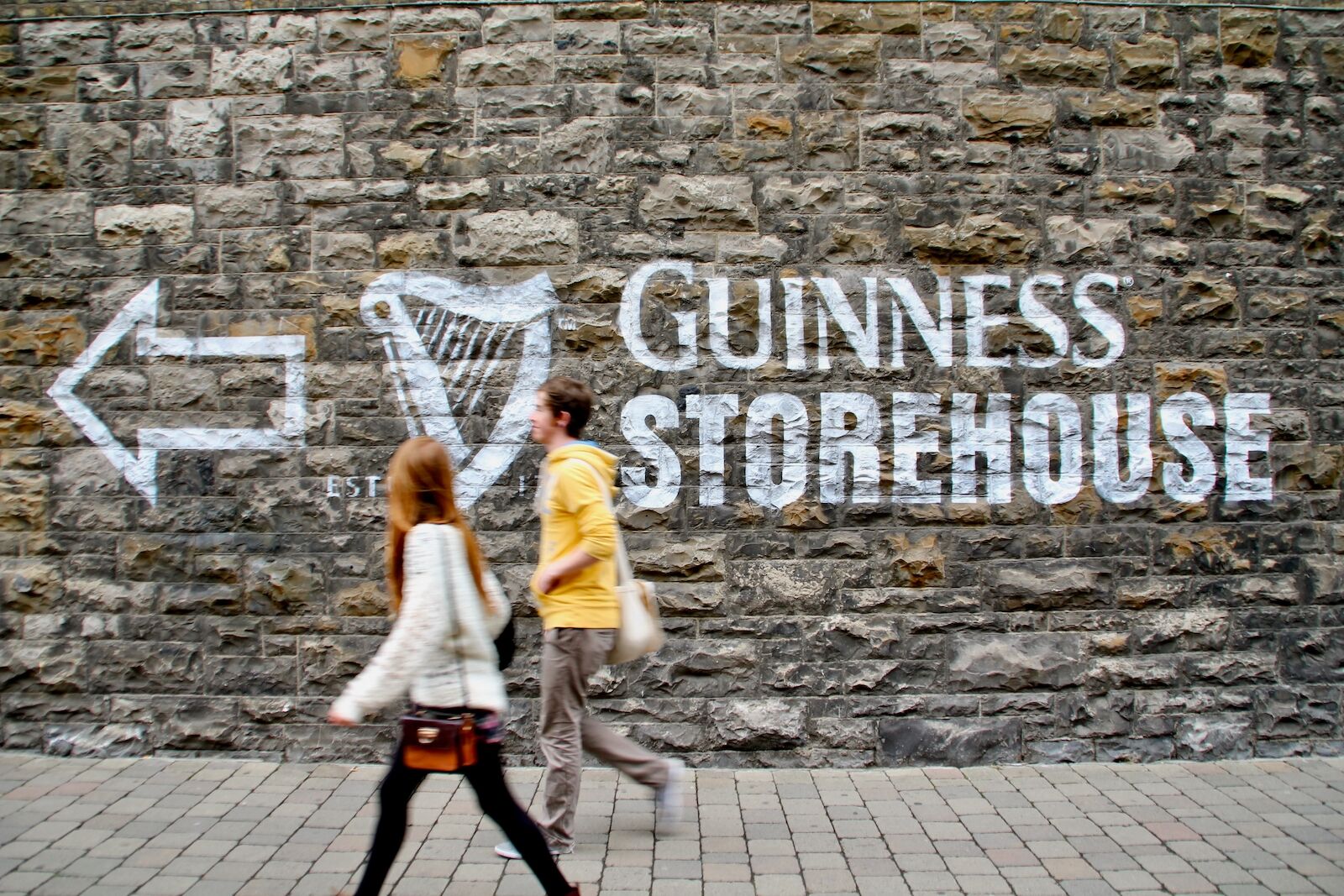 Dos jóvenes caminan junto a la señal de un muro que indica Guinness Storehouse.